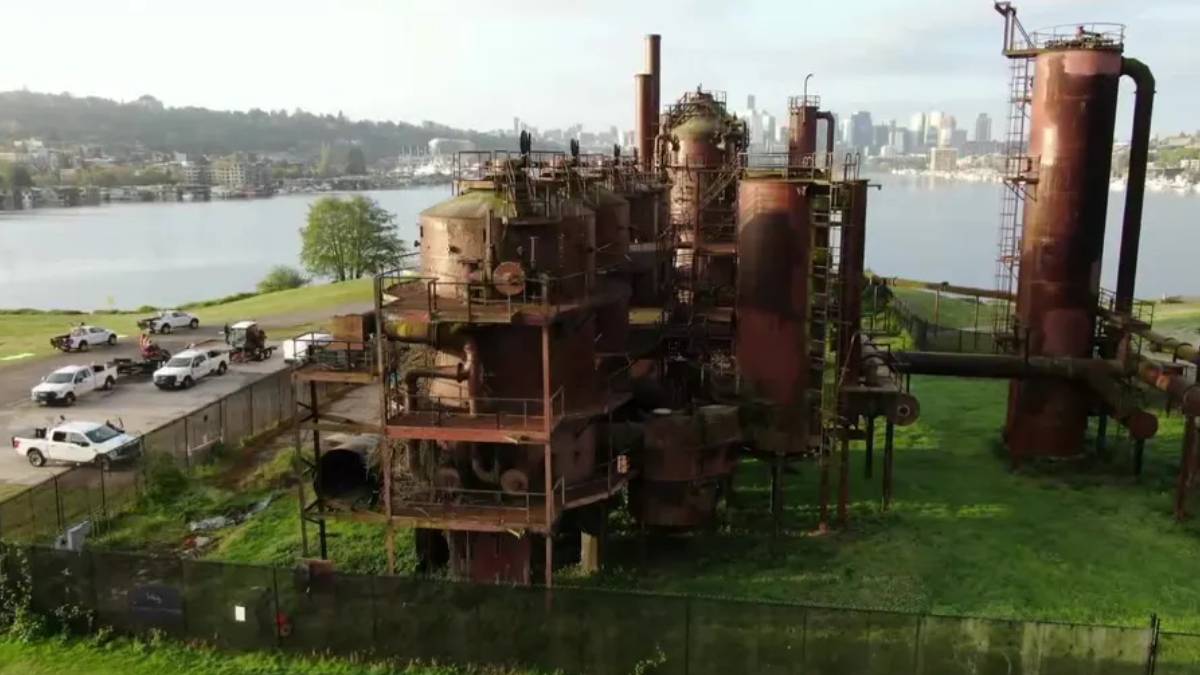 Rusty, abandoned industrial complex with tall smokestacks and interconnected tanks by a river, city skyline in the distance.