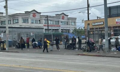 Line of people with carts and bags along a chain-link fence by a busy street, in front of a shopping center.
