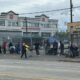Line of people with carts and bags along a chain-link fence by a busy street, in front of a shopping center.