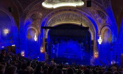 Ornate theater with carved arches and chandeliers, blue stage lighting, and an audience watching a performance from the stalls and balconies.