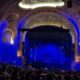 Ornate theater with carved arches and chandeliers, blue stage lighting, and an audience watching a performance from the stalls and balconies.