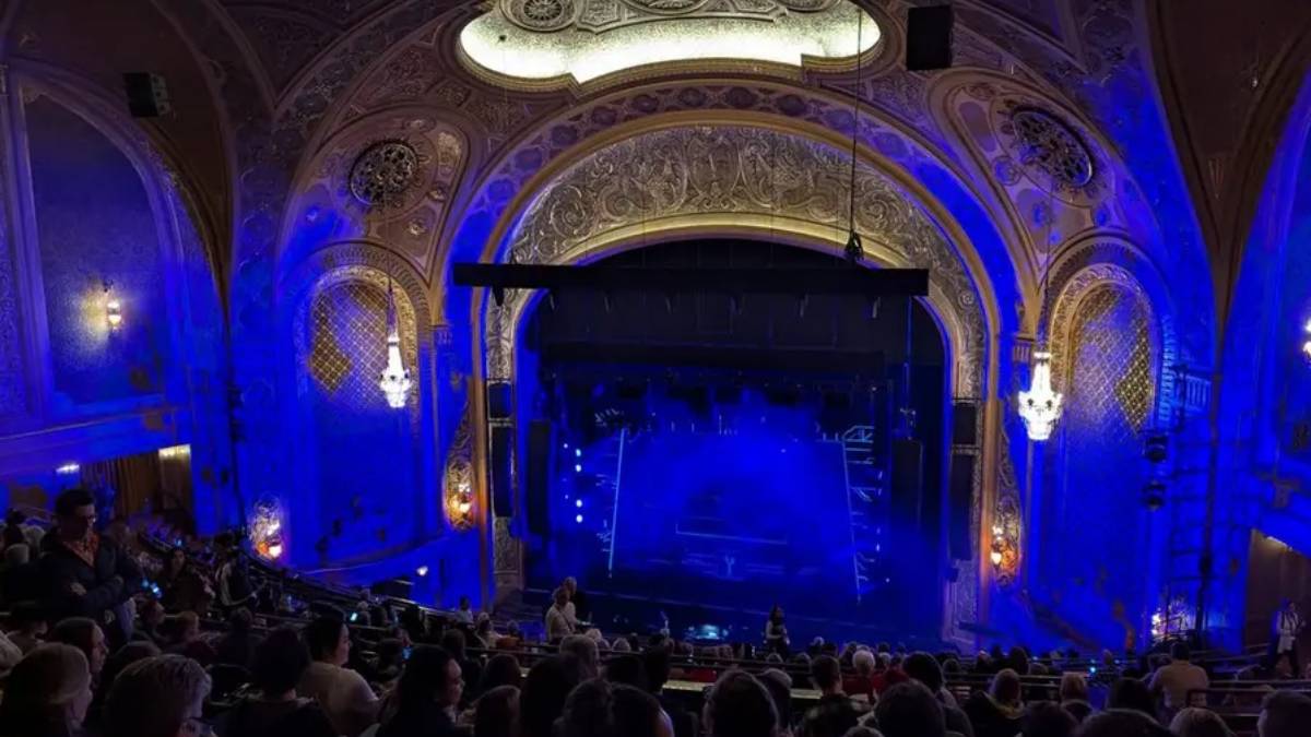 Ornate theater with carved arches and chandeliers, blue stage lighting, and an audience watching a performance from the stalls and balconies.