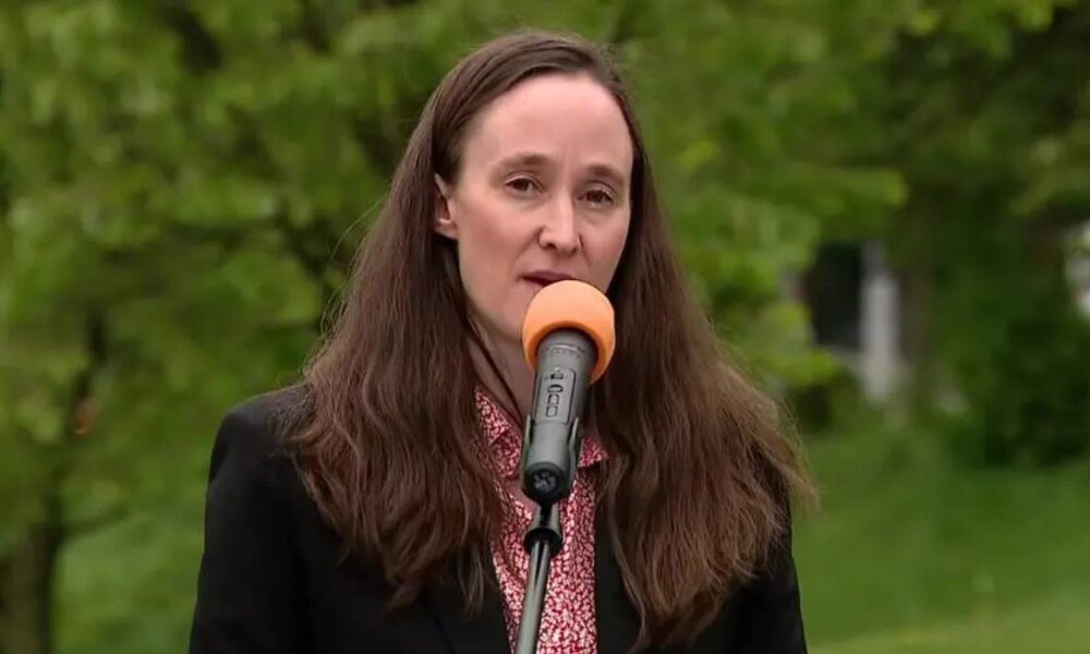 Woman in a black blazer speaks into an orange-mic windscreen outdoors, with green trees in the background.