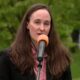 Woman in a black blazer speaks into an orange-mic windscreen outdoors, with green trees in the background.