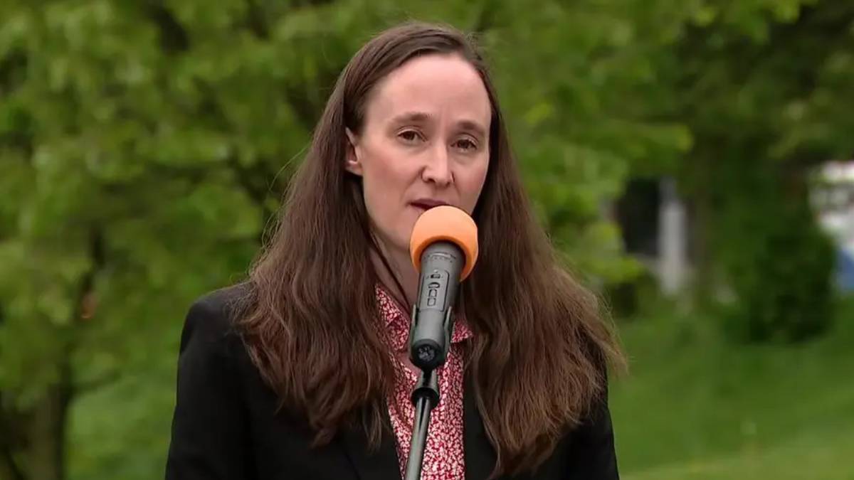 Woman in a black blazer speaks into an orange-mic windscreen outdoors, with green trees in the background.