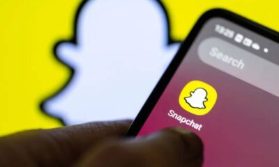 Close-up of a hand holding a smartphone displaying the Snapchat app icon on a pink gradient screen with a bright yellow background behind it.