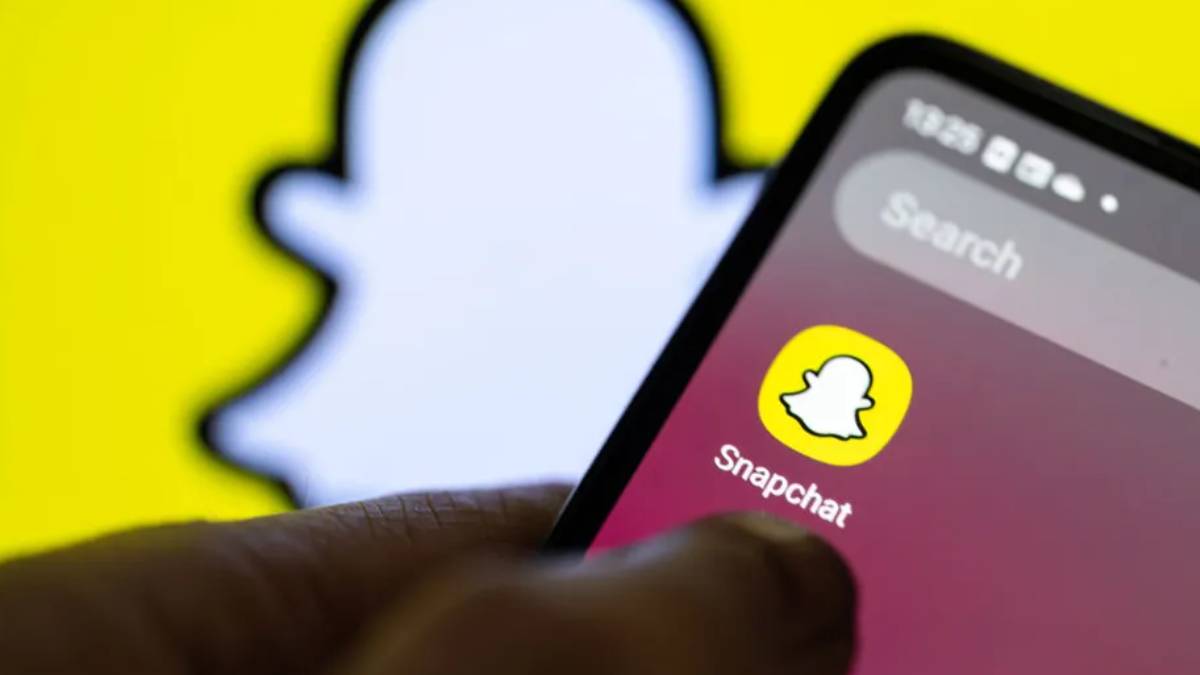 Close-up of a hand holding a smartphone displaying the Snapchat app icon on a pink gradient screen with a bright yellow background behind it.