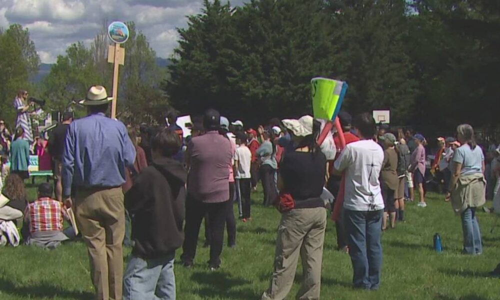 Crowd gathered in a park listening to a speaker at a small outdoor event, with signs and banners nearby.