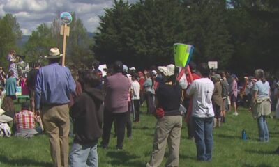 Crowd gathered in a park listening to a speaker at a small outdoor event, with signs and banners nearby.