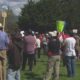 Crowd gathered in a park listening to a speaker at a small outdoor event, with signs and banners nearby.