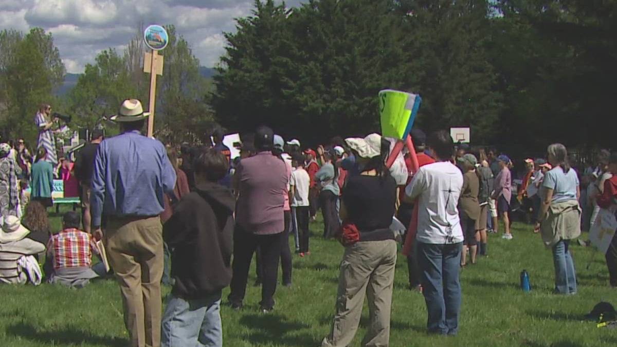 Crowd gathered in a park listening to a speaker at a small outdoor event, with signs and banners nearby.