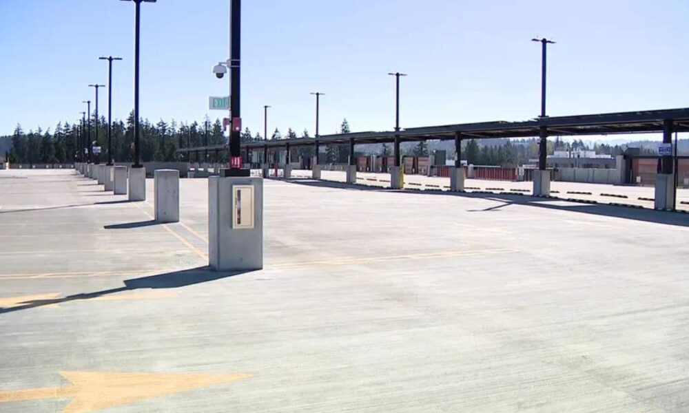 Empty parking lot with concrete dividers, tall light posts, and a long covered roadway in the distance under a clear blue sky.