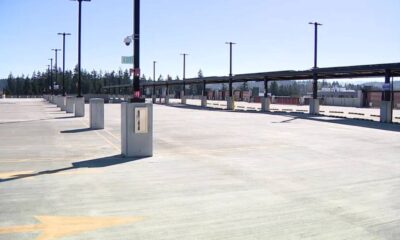 Empty parking lot with concrete dividers, tall light posts, and a long covered roadway in the distance under a clear blue sky.
