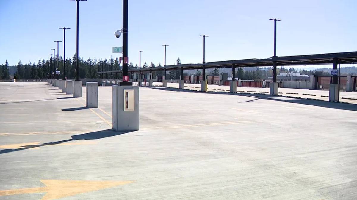 Empty parking lot with concrete dividers, tall light posts, and a long covered roadway in the distance under a clear blue sky.