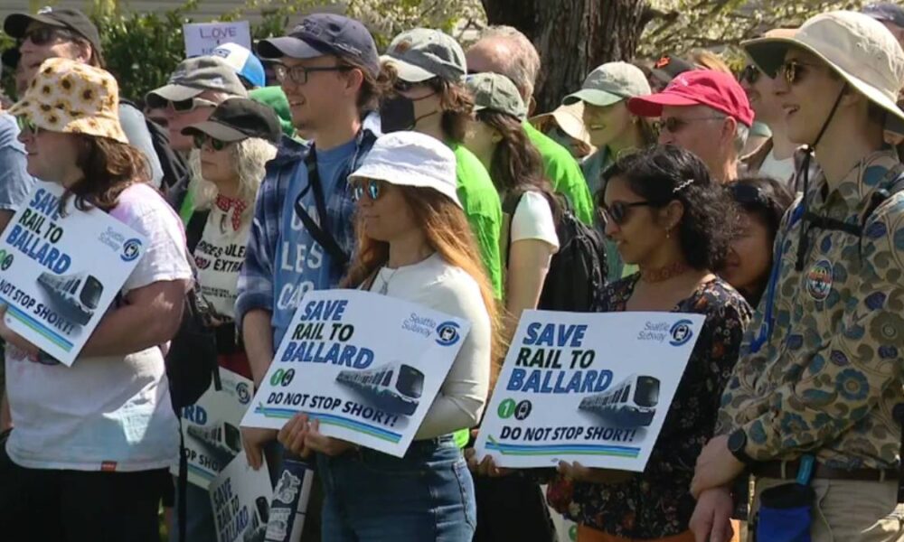 Group of diverse protesters at a rally, holding signs that say 'Save Rail to Ballard' and 'Do not stop short!'