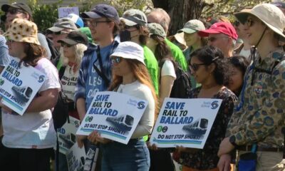 Group of diverse protesters at a rally, holding signs that say 'Save Rail to Ballard' and 'Do not stop short!'