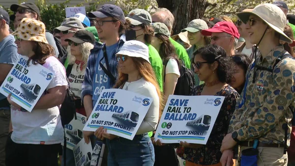 Group of diverse protesters at a rally, holding signs that say 'Save Rail to Ballard' and 'Do not stop short!'