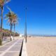 Palm-lined boardwalk with a sandy beach and calm blue sea under a clear sky; people strolling in the distance.