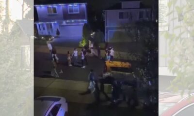 Group of people walking along a residential street at night, with houses and string lights visible in the background.