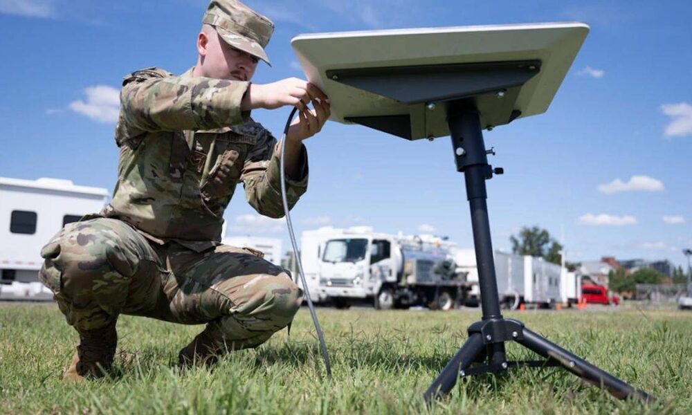 Soldier in camouflage kneels on grass, connecting cables to a portable satellite dish on a tripod outdoors with support trucks in the background.