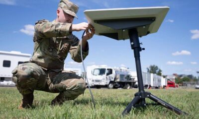 Soldier in camouflage kneels on grass, connecting cables to a portable satellite dish on a tripod outdoors with support trucks in the background.
