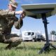 Soldier in camouflage kneels on grass, connecting cables to a portable satellite dish on a tripod outdoors with support trucks in the background.