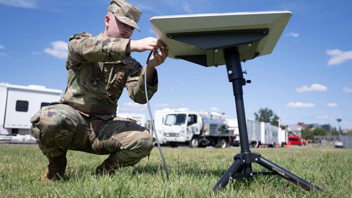 Soldier in camouflage kneels on grass, connecting cables to a portable satellite dish on a tripod outdoors with support trucks in the background.