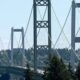 Suspension bridge with tall towers and crisscrossing cables over a forested valley, cars on the roadway beneath.
