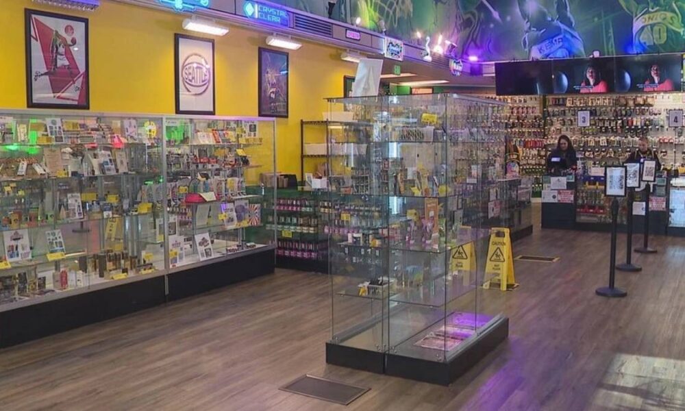Bright retail store interior with glass display cases along the walls and items for sale; two staff members stand near the back counter.