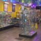 Bright retail store interior with glass display cases along the walls and items for sale; two staff members stand near the back counter.
