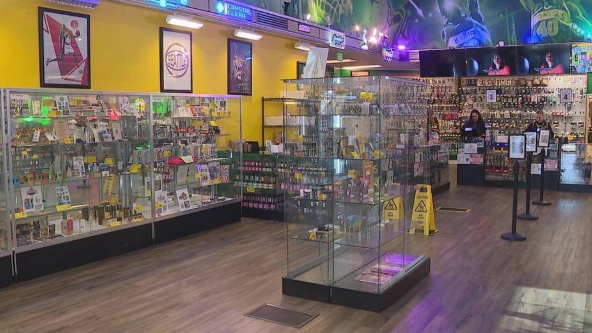 Bright retail store interior with glass display cases along the walls and items for sale; two staff members stand near the back counter.