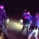 Nighttime roadside police scene with several officers surrounding a person near a white vehicle, illuminated by flashing emergency lights and a dark backdrop of the road.