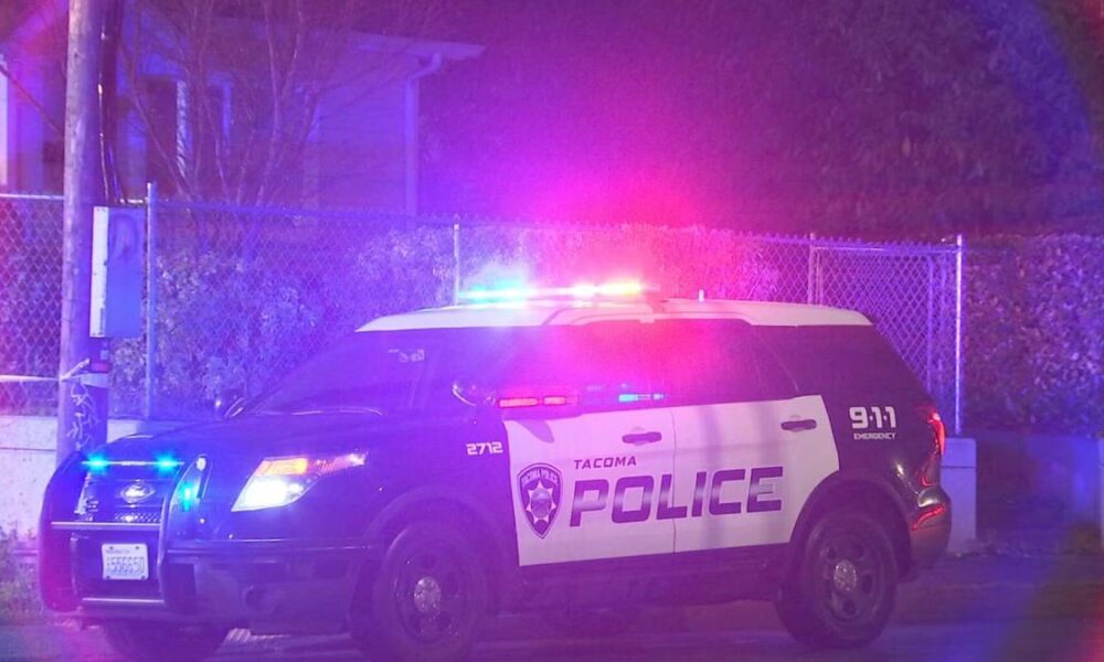 Tacoma Police Department SUV with red and blue emergency lights flashing at night beside a chain-link fence.