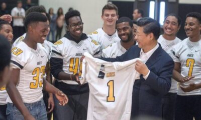 Team of football players in white jerseys celebrate as a man in a suit presents a new jersey marked number 1.