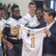 Team of football players in white jerseys celebrate as a man in a suit presents a new jersey marked number 1.