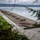 Lakeside park with a concrete path, logs on the pebble beach, and picnic tables under a cloudy sky.