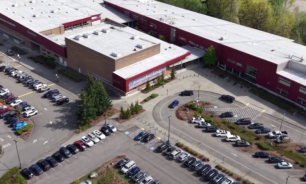 Aerial view of a large red-brick school building with a white roof, surrounded by parking lots and a circular driveway with cars and landscaped medians.