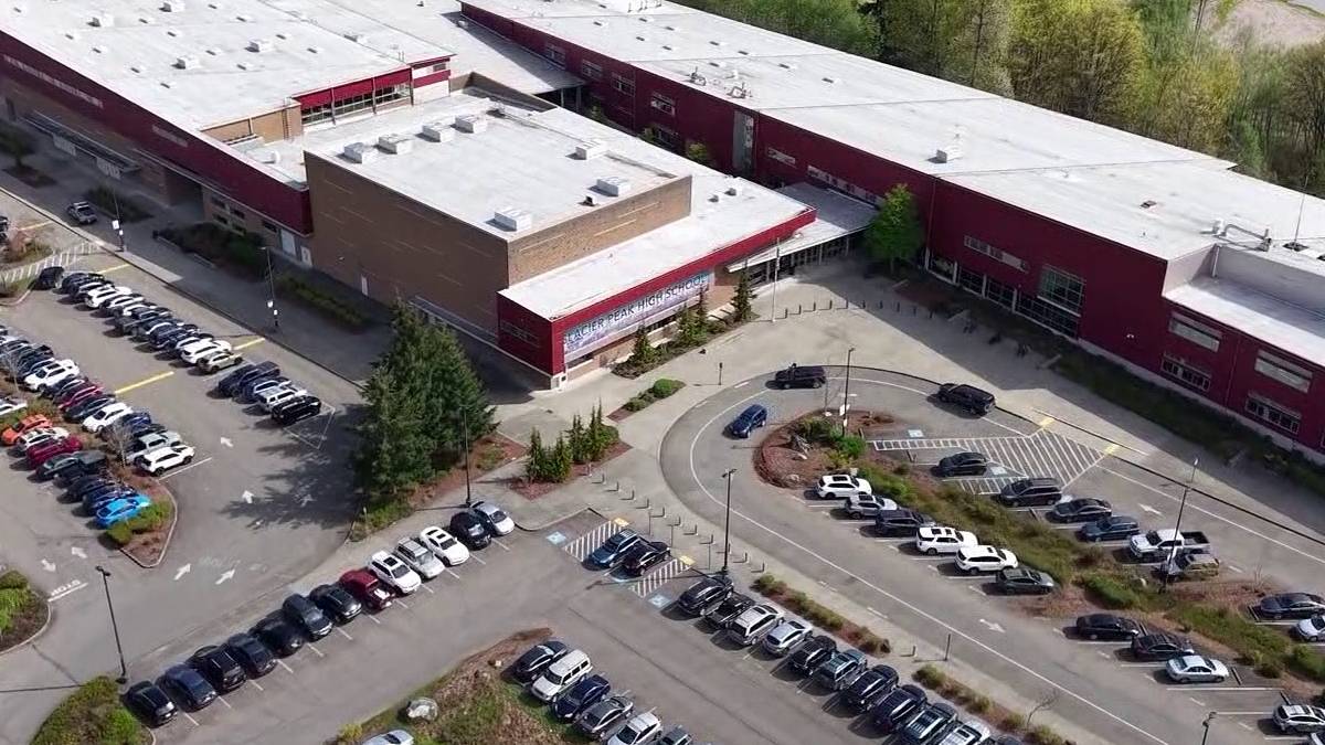 Aerial view of a large red-brick school building with a white roof, surrounded by parking lots and a circular driveway with cars and landscaped medians.