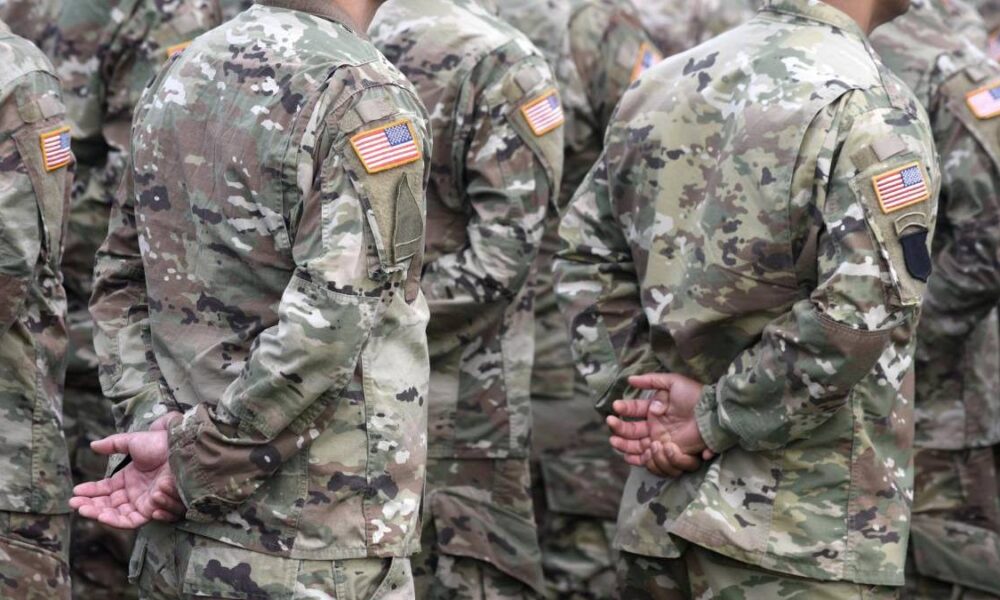 Group of soldiers in camouflage uniforms standing in formation with hands behind their backs, US flag patches on sleeves.