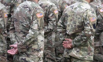 Group of soldiers in camouflage uniforms standing in formation with hands behind their backs, US flag patches on sleeves.