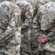 Group of soldiers in camouflage uniforms standing in formation with hands behind their backs, US flag patches on sleeves.