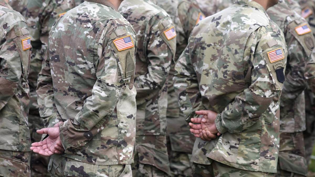 Group of soldiers in camouflage uniforms standing in formation with hands behind their backs, US flag patches on sleeves.