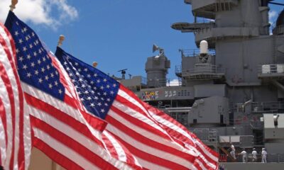 American flags waving in foreground with a gray naval warship and sailors in white uniforms in the background