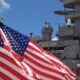 American flags waving in foreground with a gray naval warship and sailors in white uniforms in the background