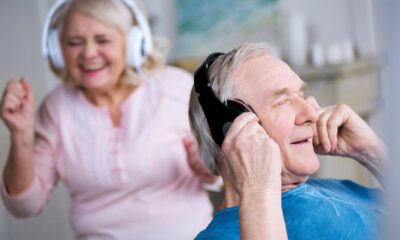 Two smiling seniors wear wireless over-ear headphones, the man in the foreground adjusting his headphones while the woman in the background dances along to music.