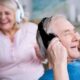 Two smiling seniors wear wireless over-ear headphones, the man in the foreground adjusting his headphones while the woman in the background dances along to music.