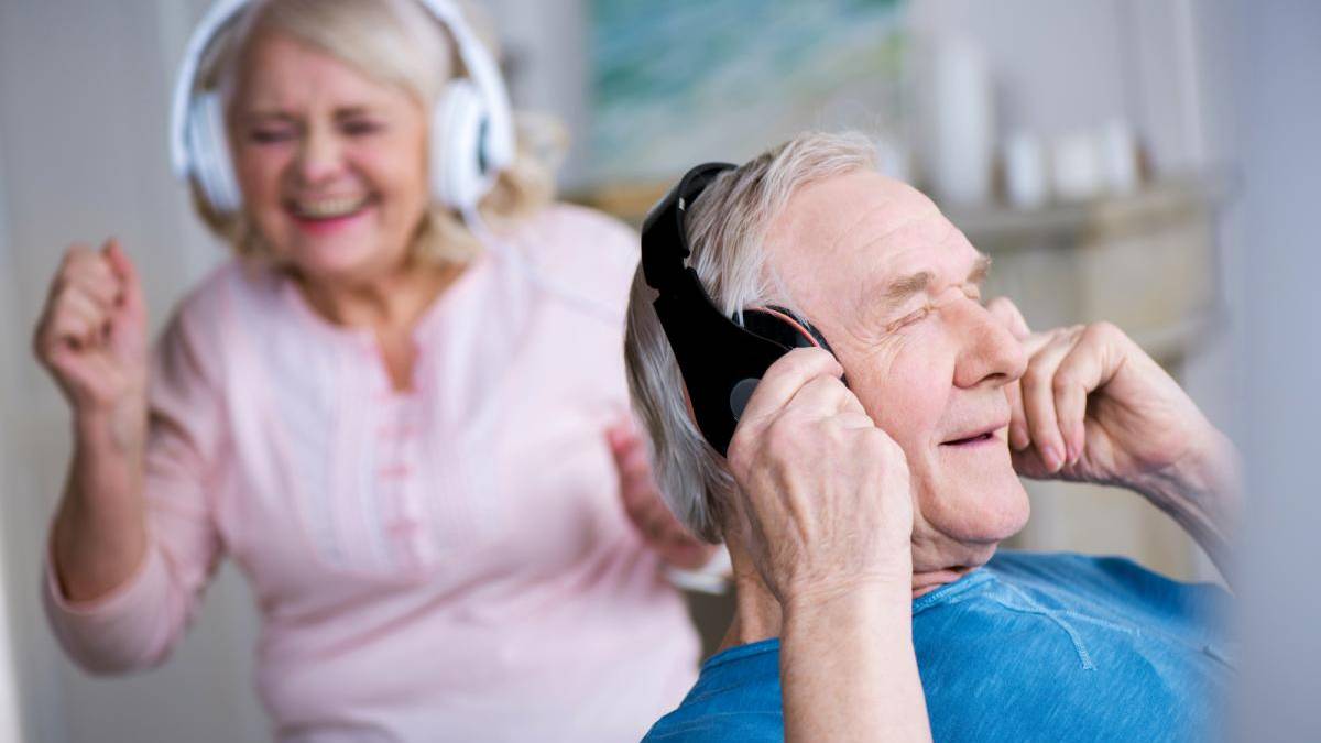 Two smiling seniors wear wireless over-ear headphones, the man in the foreground adjusting his headphones while the woman in the background dances along to music.