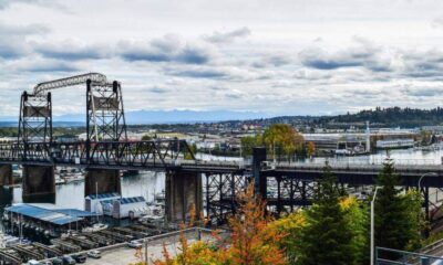 Steel lift bridge over a harbor with boats, cityscape in distance and autumn trees in the foreground.