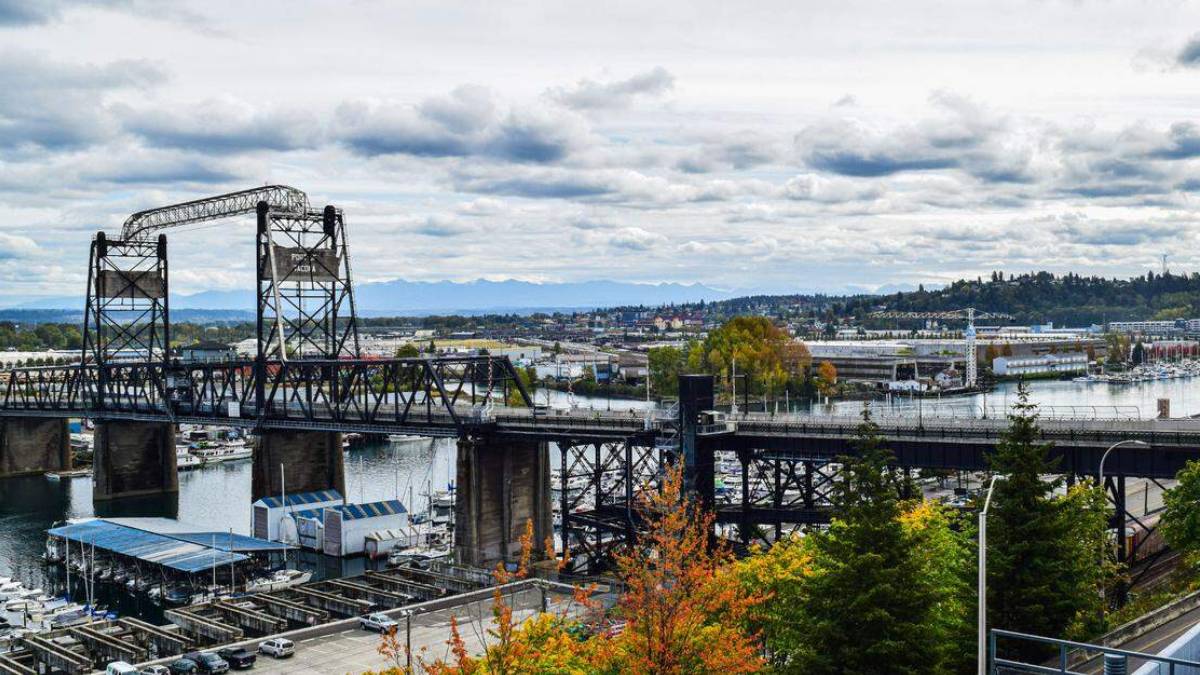 Steel lift bridge over a harbor with boats, cityscape in distance and autumn trees in the foreground.