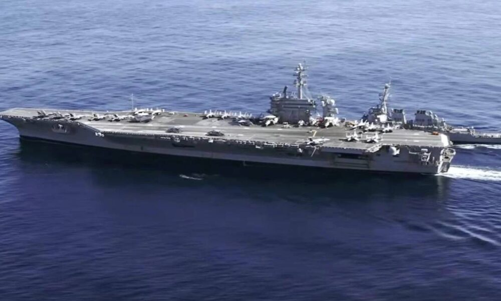 Large aircraft carrier at sea with fighter jets lined up on the deck and a smaller escort ship nearby in blue water.